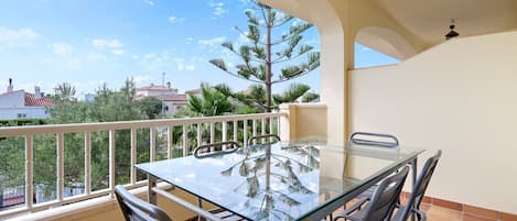 A balcony with a glass table and chairs, a white railing, and a view of trees and buildings.