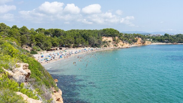 A coastal beach with clear waters, a sandy shore, and a backdrop of cliffs and greenery.
