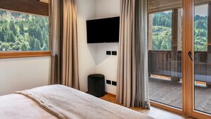 A bedroom with a bed, a television mounted on the wall, a small stool, and a sliding glass door leading to a balcony with a forest view. - Villa Sofia Apartment 'I' with Mountain View, Private Terrace and Wi-Fi (Selva di Val Gardena)