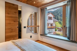 Une chambre à coucher avec un lit, une porte en bois, une grande fenêtre avec des rideaux et une vue sur une maison et des montagnes.