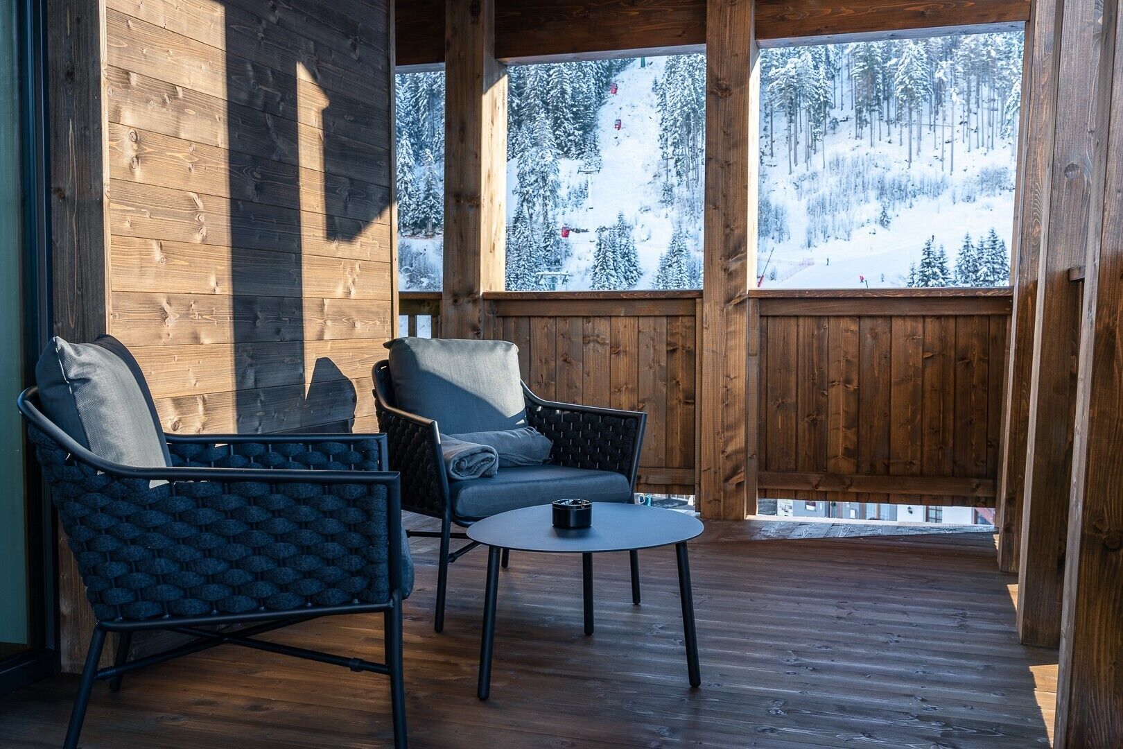 Eine Holzterrasse mit zwei Stühlen und einem kleinen Tisch mit Blick auf einen verschneiten Wald.