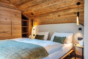 A wooden ceiling, a bed with white bedding, a bedside table with a lamp, and a shelf unit.