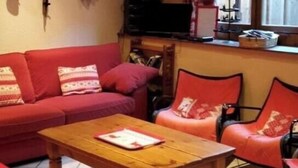 Ein Wohnzimmer mit einem roten Sofa, einem Couchtisch aus Holz und einem roten Sessel. Ein Fernseher steht auf einem Schrank, ein Fenster mit Vorhängen und eine Wandregal mit Skiern und anderen Gegenständen.