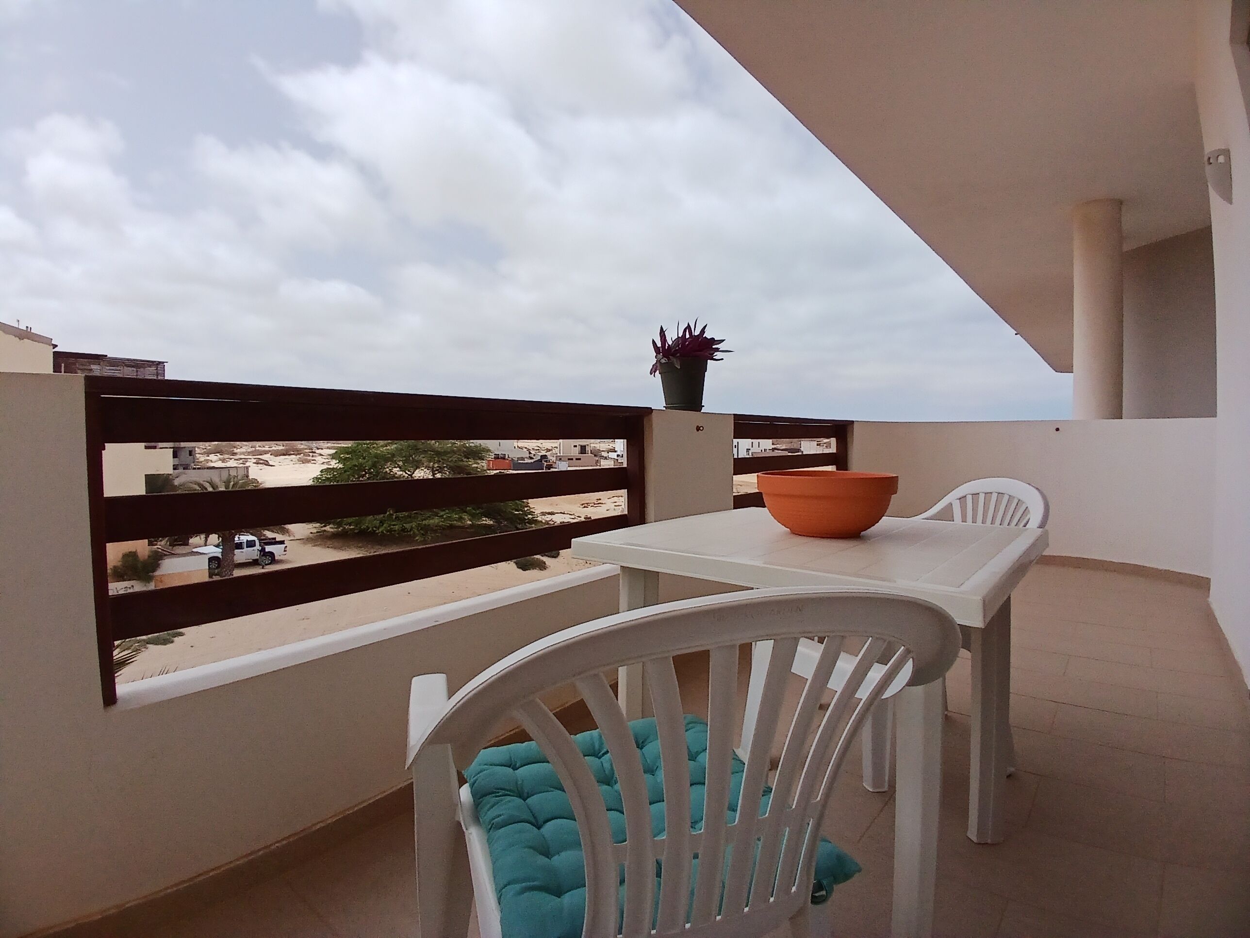 Un balcón con mesa y silla blancos, una planta en maceta y vistas del entorno.
