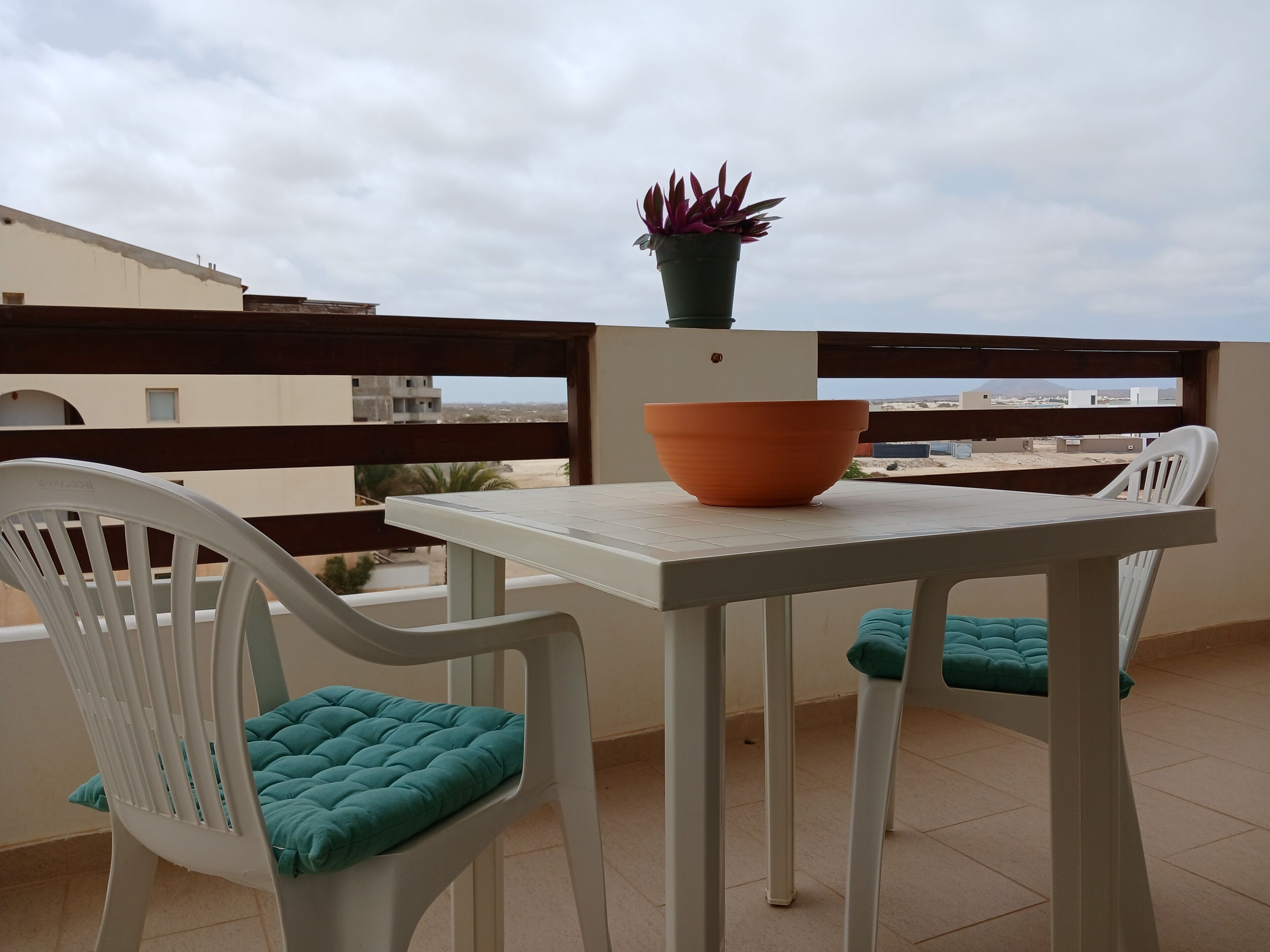 A balcony with a white table, two white chairs with teal cushions, a terracotta bowl, and a potted plant.