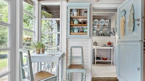 A rustic kitchen with a wooden table, a blue chair, and a cabinet with dishes and jars.