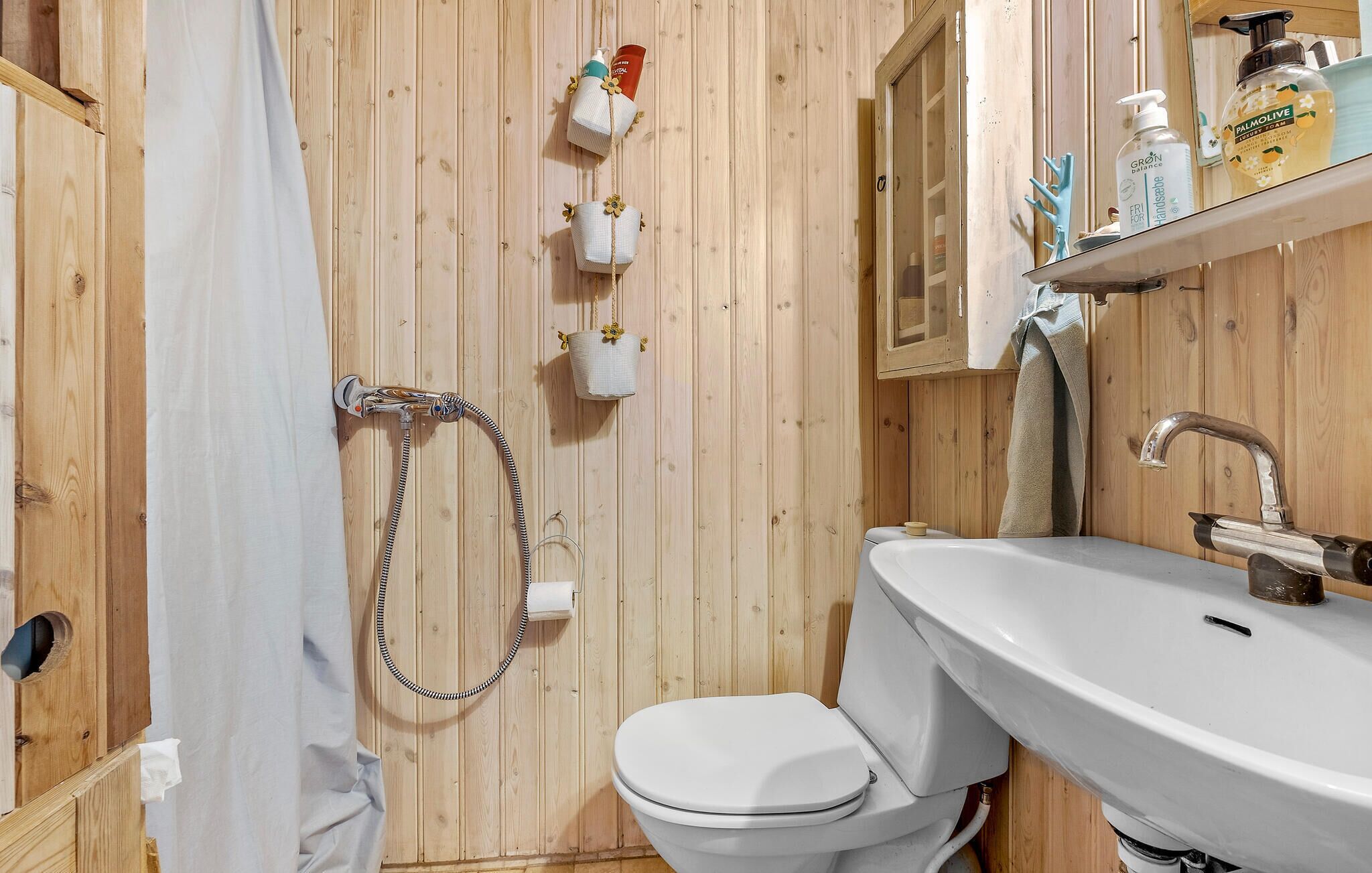 A wooden-paneled bathroom with a white toilet, a shower with a hose, and a sink with toiletries.