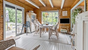 A cozy cabin interior with a wooden ceiling, a dining area with wicker chairs, a large window, and a television.