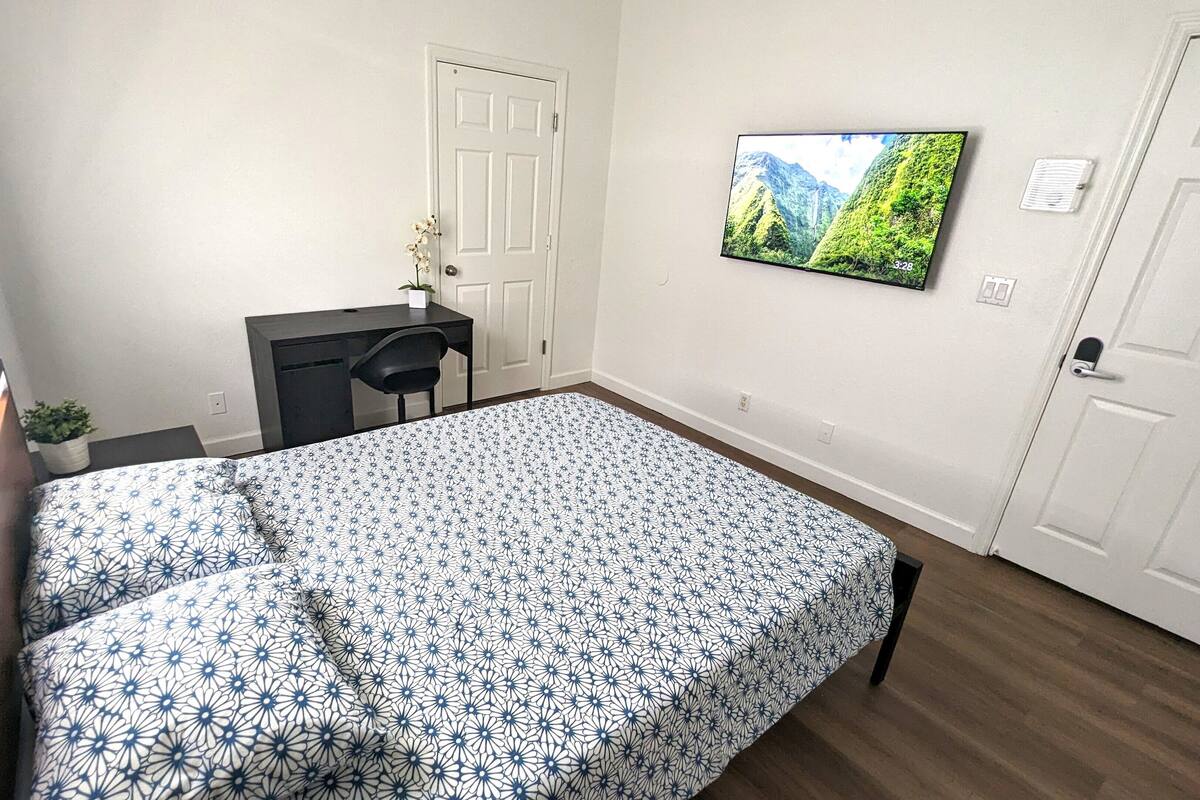 Un dormitorio con cama, escritorio, silla, planta en maceta, televisor y una puerta.