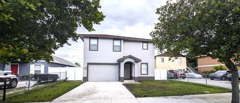 A two-story residential building with a garage, surrounded by a fenced yard and parked cars.