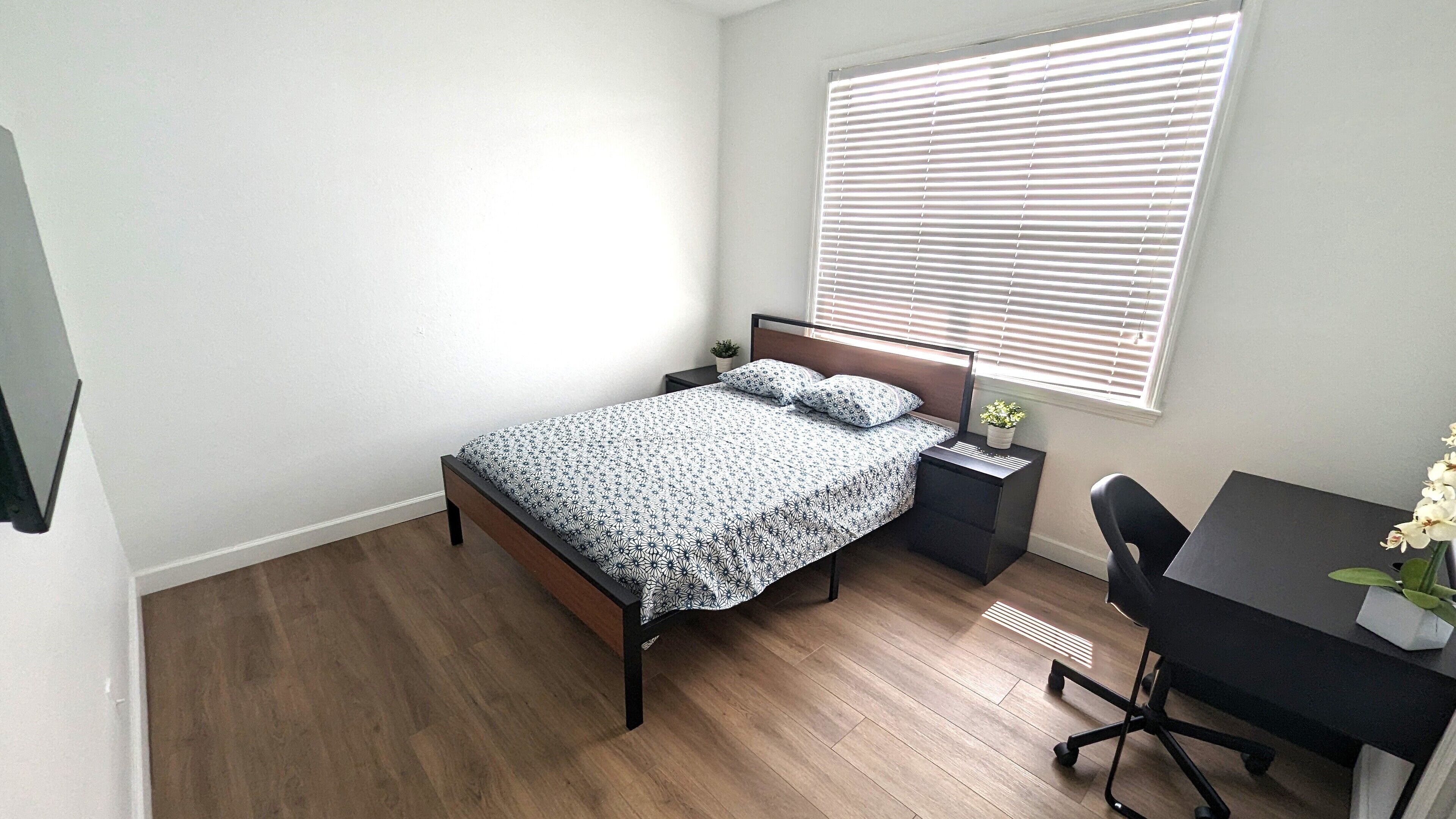 A bedroom with a bed, a desk, and a window with blinds.