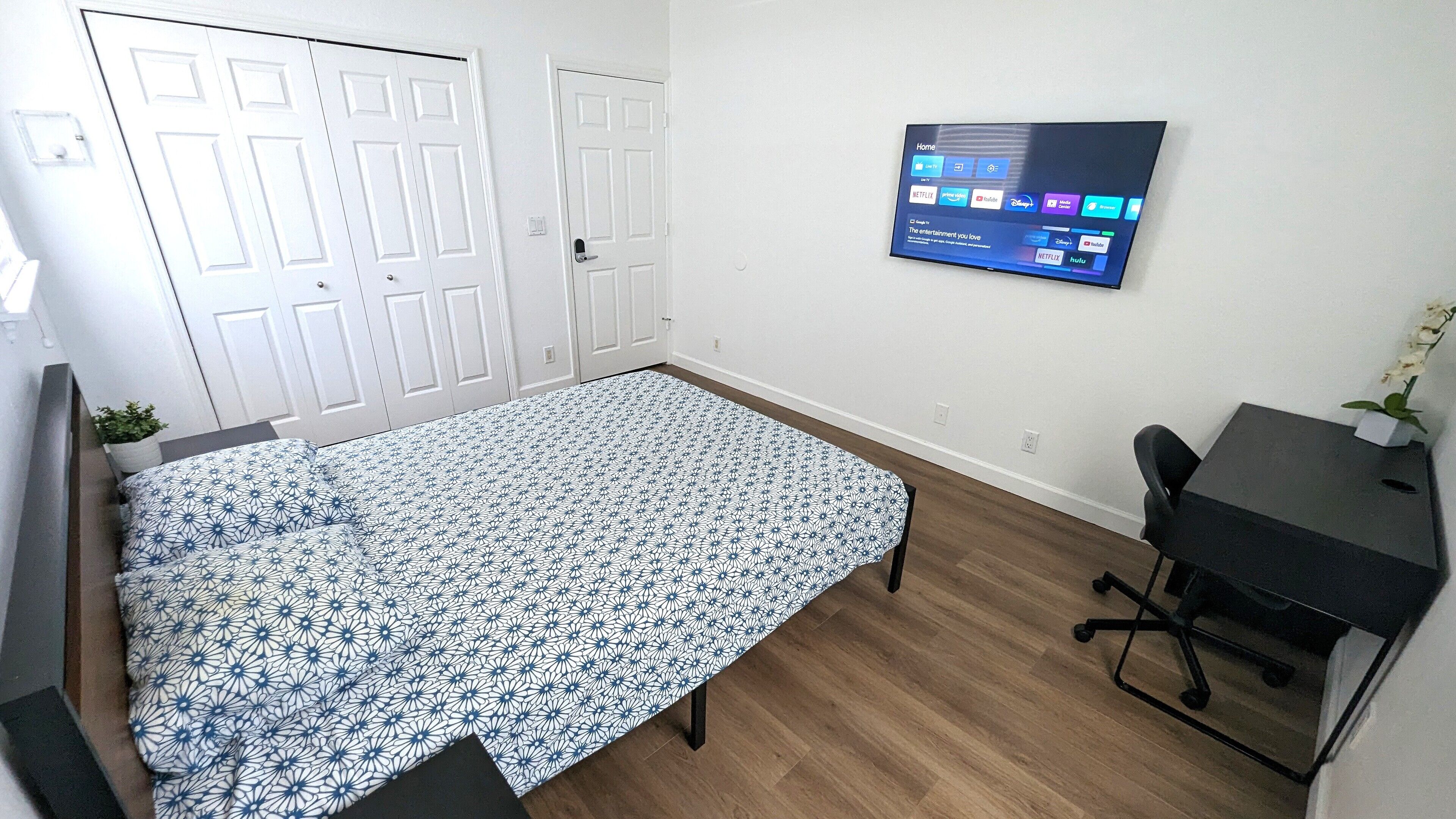 A bedroom with a bed, a desk, and a TV mounted on the wall displaying streaming services.
