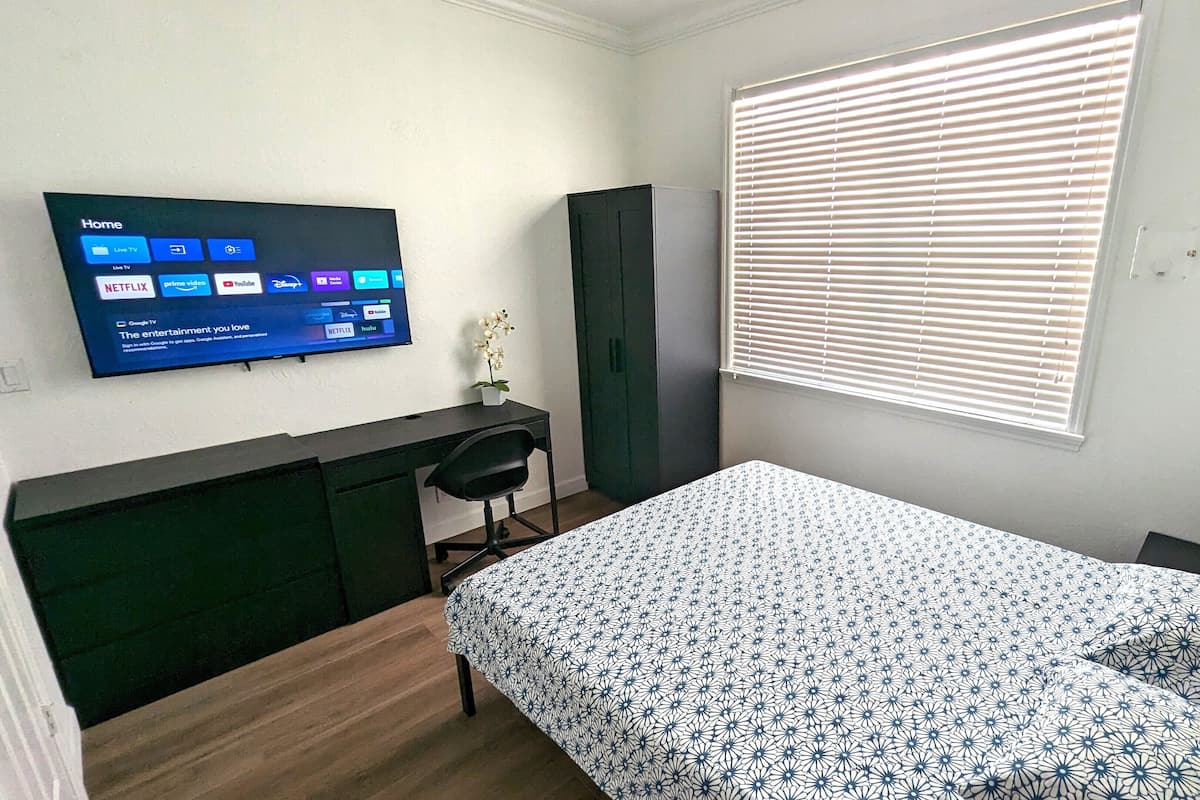 Un dormitorio con cama, escritorio, silla, televisor montado en la pared que muestra servicios de transmisión y una ventana con persianas.
