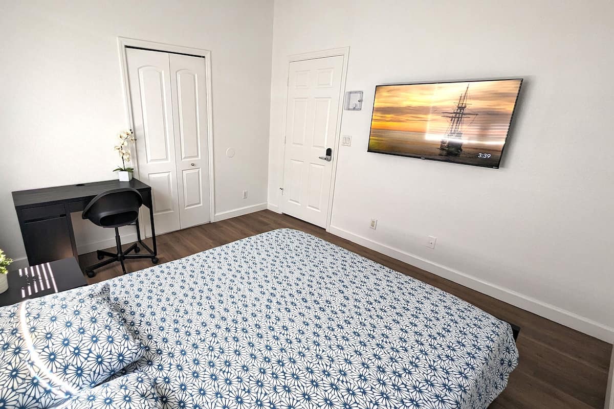 Un dormitorio con una cama, un escritorio, dos puertas, una planta en maceta y un televisor en la pared.
