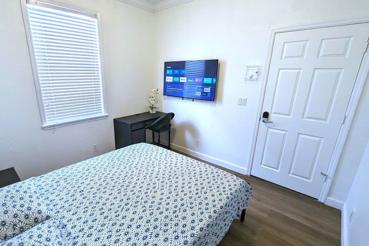 Un dormitorio con cama, escritorio, planta en maceta, televisor en la pared y una puerta.