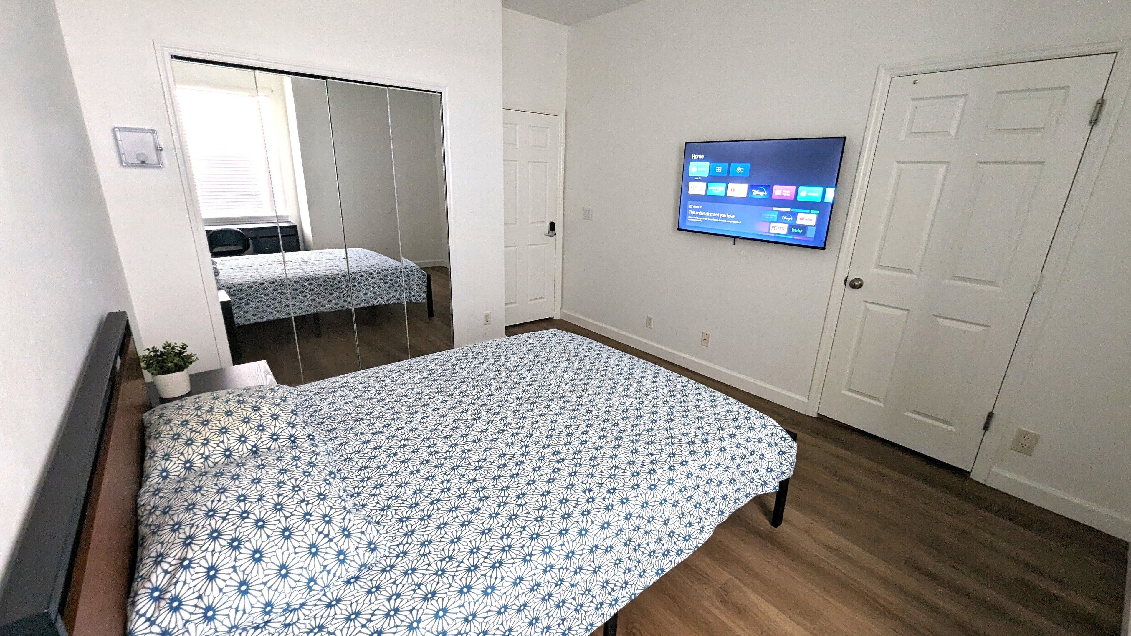 A bedroom with a bed, a TV mounted on the wall, a door, and a window with blinds.