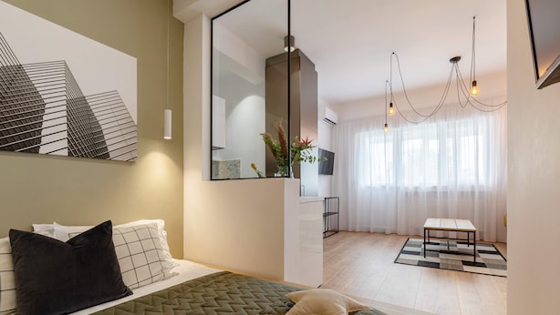 A modern bedroom with a bed, a large mirror, a hanging light fixture, and a view of a cityscape through sheer curtains.