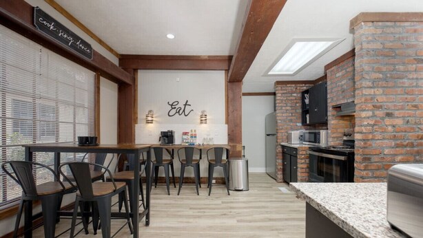 Una cocina moderna con pared de ladrillo, vigas de madera, zona de comedor con sillas y un cartel que dice 'cook sing dance'.