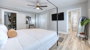 Un dormitorio con cama, ventilador de techo, televisor, espejo y planta.
