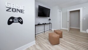 Una sala de estar moderna con un letrero de zona de juegos, un televisor de pantalla plana montado en una pared con diseño de ladrillo y dos otomanas marrones en el suelo.