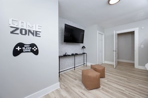 A modern living room with a gaming zone sign, a flat-screen TV mounted on a brick-patterned wall, and two brown ottomans on the floor.