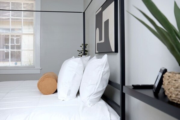 A bed with white bedding, a brown pillow, and two white pillows. There is a framed artwork on the wall and a plant on the right.