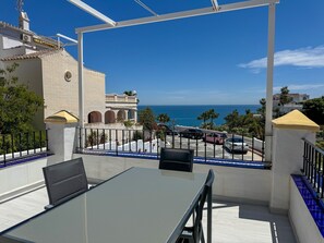 A balcony with a table and chairs, overlooking a street with parked cars and a view of the sea.