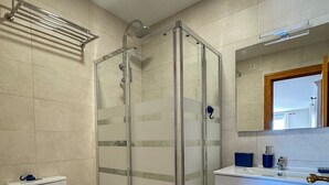 Kylpyhuoneessa on lasiseinällä varustettu suihku, wc, peilillä varustettu pesuallas ja sininen matto lattialla.