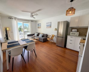 A modern living room with a sofa, dining table, chairs, a ceiling fan, and a kitchen area with a refrigerator and microwave.