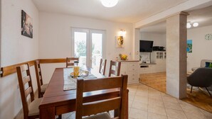 A dining area with a wooden table and chairs, a TV in the background, and a kitchen area visible through an open door. - Amazing home in Prolozac Donji (Prolozac Donji)
