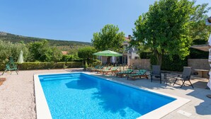 A rectangular swimming pool with clear blue water, surrounded by lounge chairs and umbrellas, set against a backdrop of lush greenery and a mountainous landscape. - Amazing home in Prolozac Donji (Prolozac Donji)