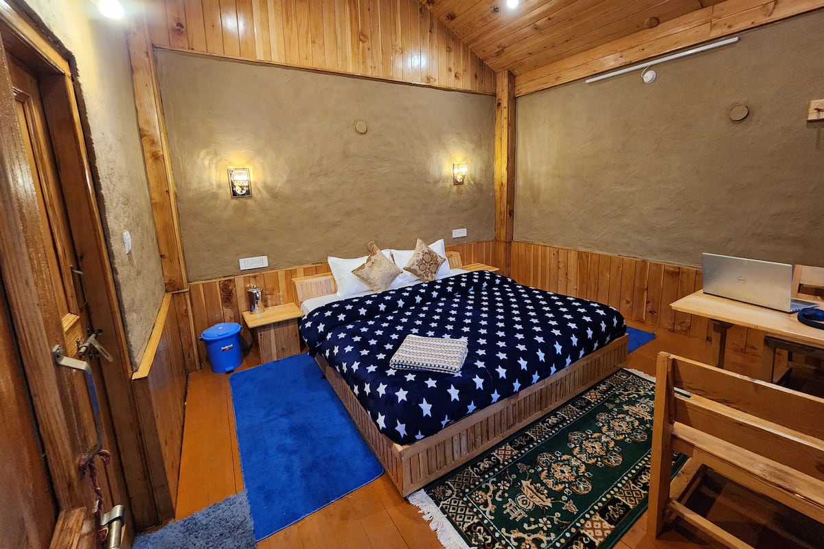 A wooden-paneled room with a bed, a desk with a laptop, and a small table with a lamp.