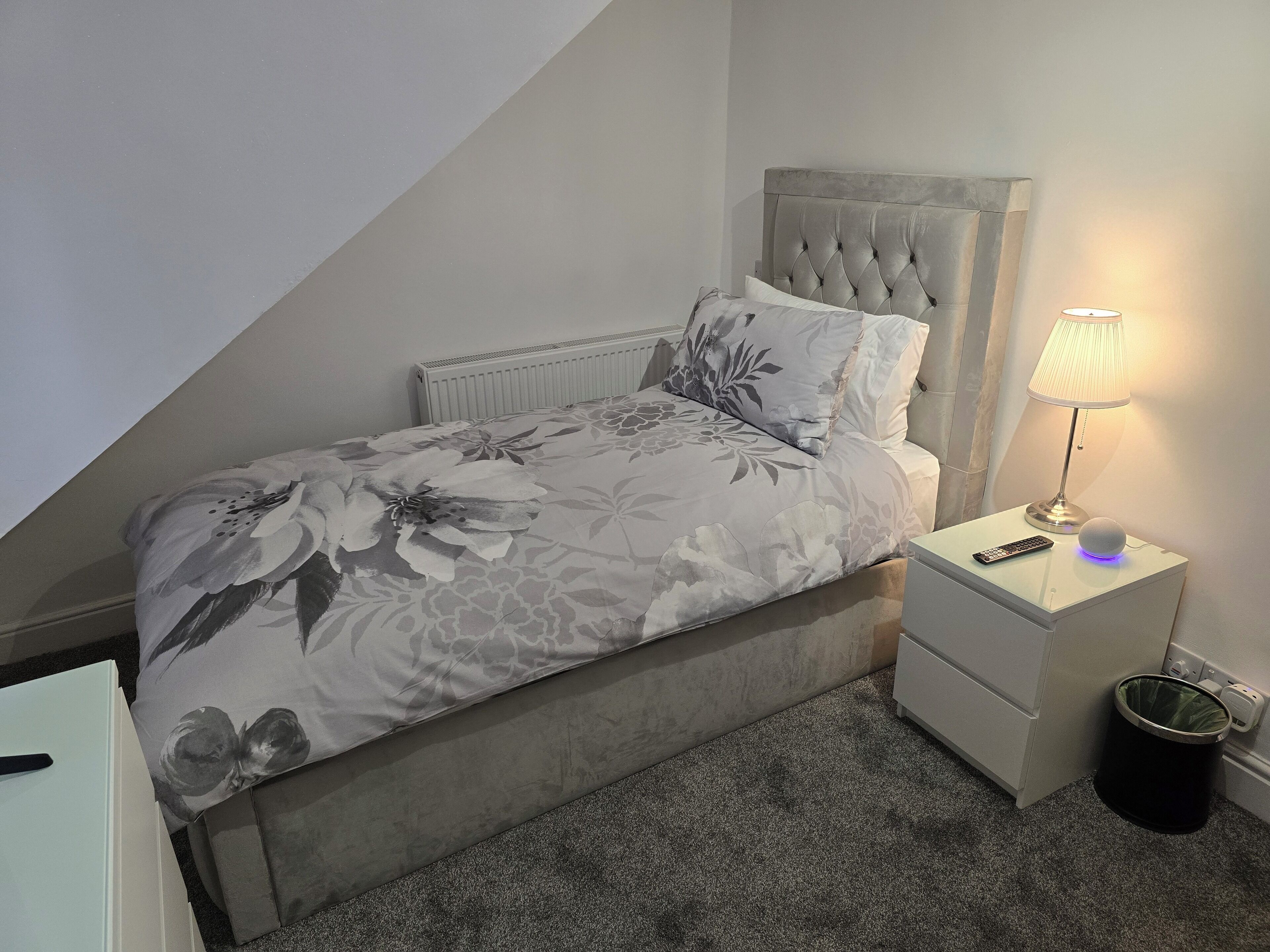 A bedroom with a bed, a nightstand, a lamp, and a trash can.