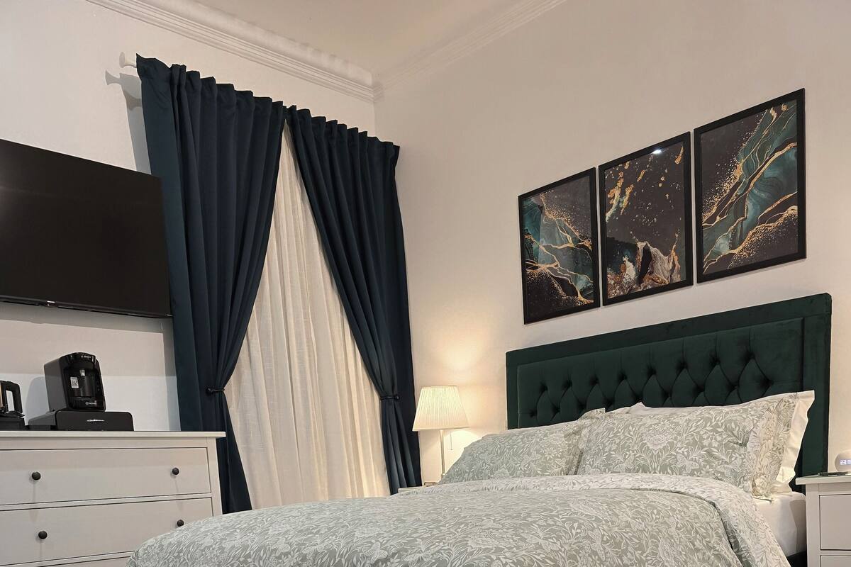 A neatly arranged bedroom with a tufted headboard, a bed with floral bedding, a bedside table, a lamp, and artwork on the wall.
