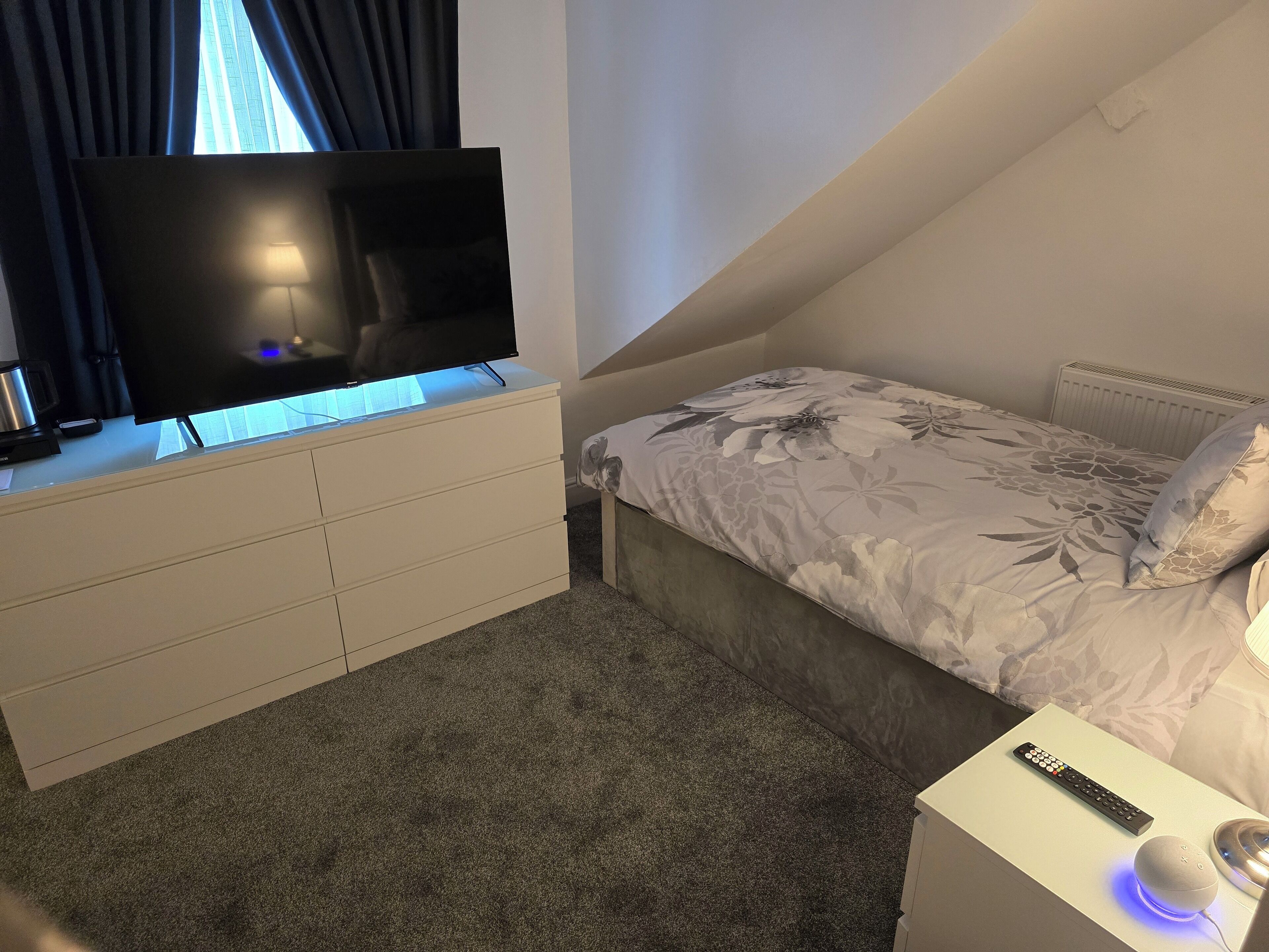 Un dormitorio con una televisión sobre un tocador, una cama con ropa de cama floral, un control remoto y un altavoz inteligente en una mesita de noche.