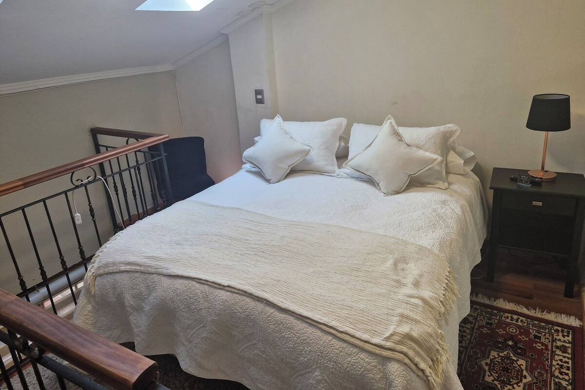 A bedroom with a bed, pillows, a nightstand, a lamp, a remote control, a wooden railing, and a skylight.