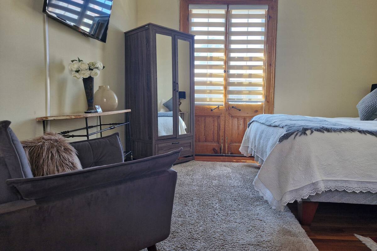 A bedroom with a bed, a chair, a wardrobe, a mirror, and a window with blinds.