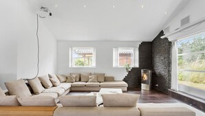 A modern living room with a large sectional sofa, a fireplace, and large windows offering a view of the outdoors. - 3 bedroom amazing home in Rødby (Rødby)