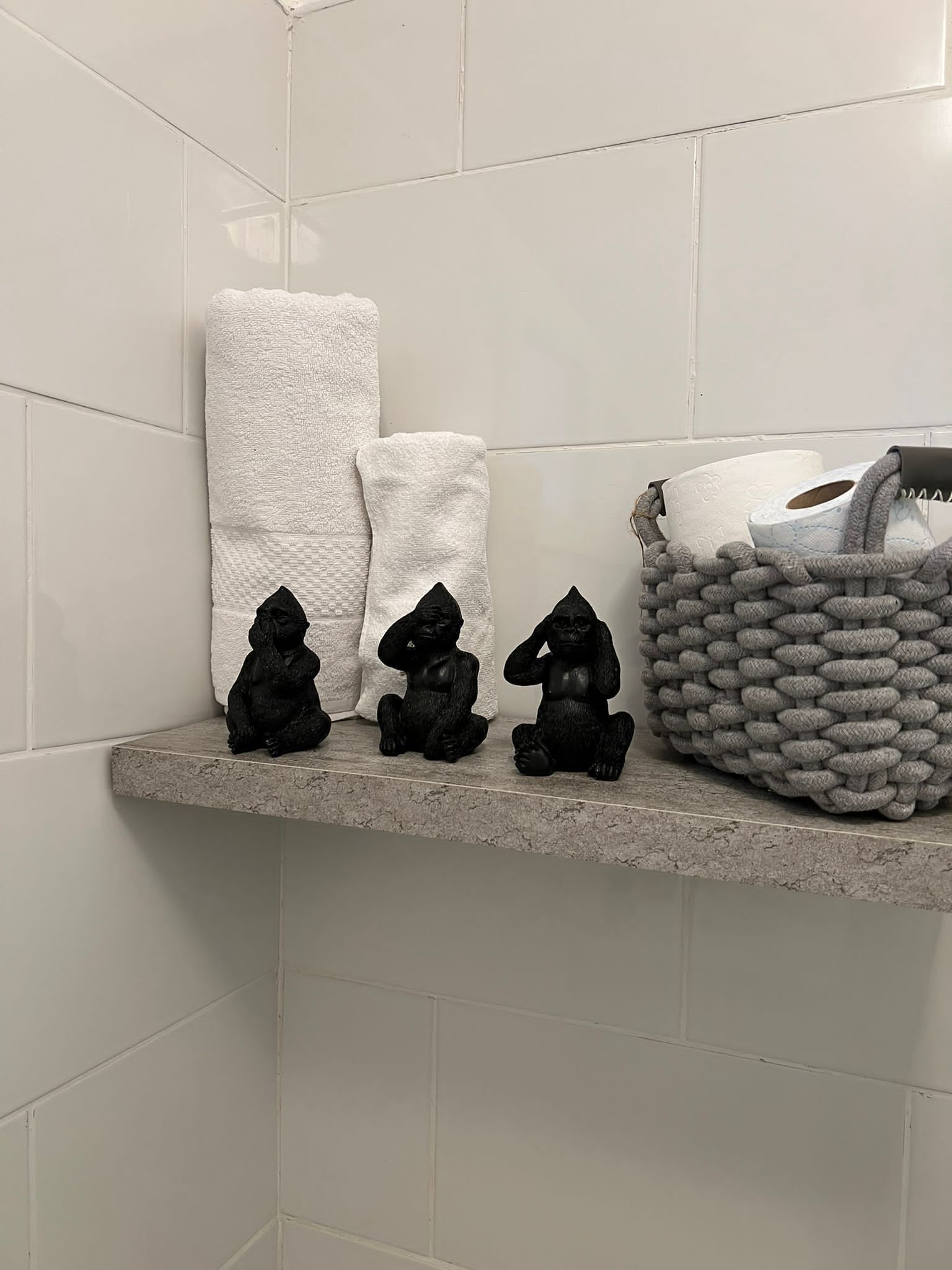 Ein Badezimmer mit weißen Fliesen, einem Regal mit drei schwarzen Figuren, zwei weißen Handtüchern und einem grauen Korbwäsches mit Toilettenartikeln.