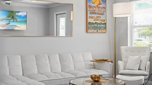 Una sala de estar moderna con un sofá blanco, una mesa de centro de vidrio, un sillón blanco y un cartel grande con una escena de playa.