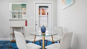 Una habitación con una mesa de vidrio rodeada de cuatro sillas blancas, un jarrón azul, un extintor y una puerta con un panel de vidrio decorativo.