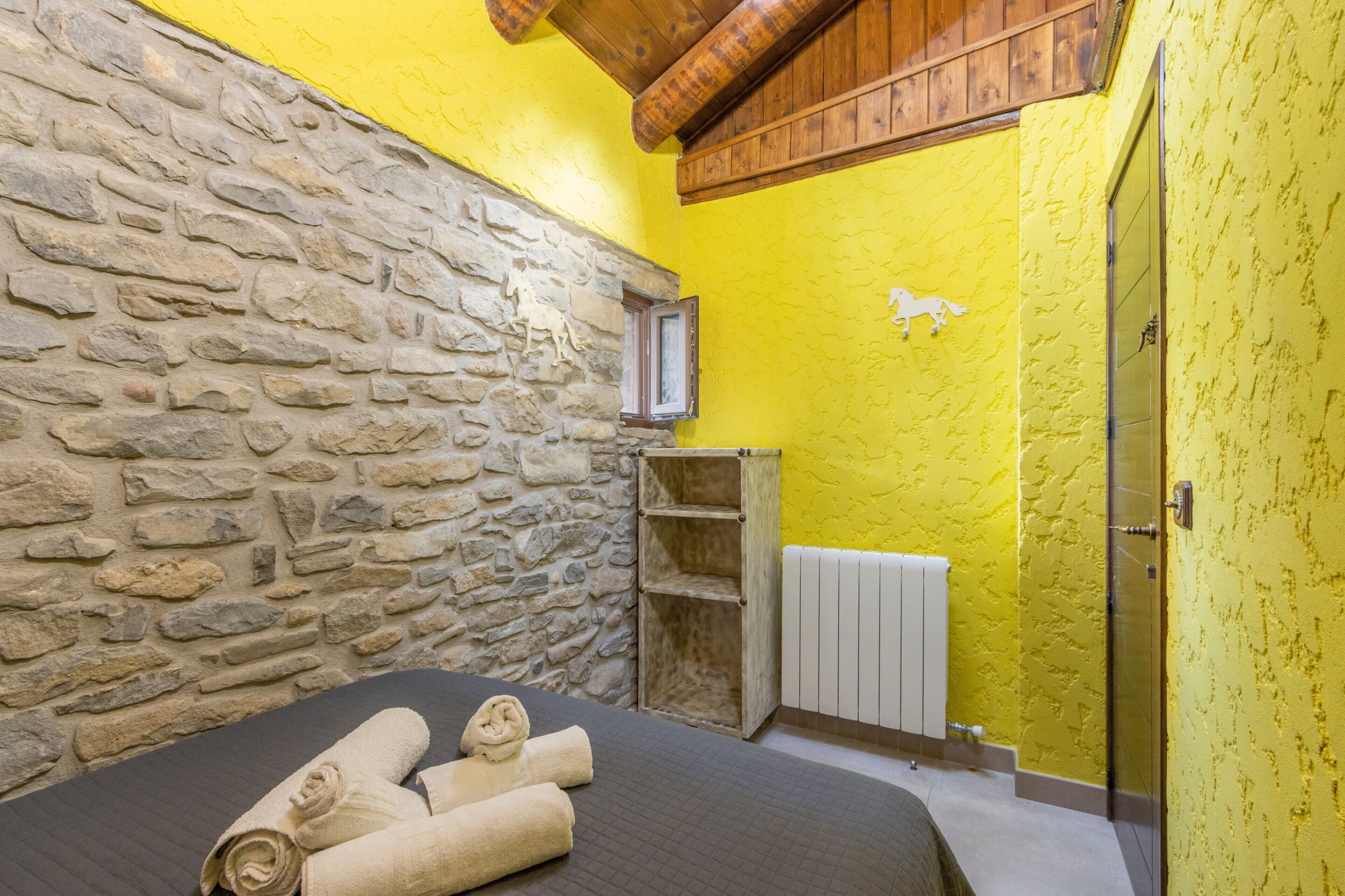 A room with a stone wall, a wooden shelf, a bed with rolled towels, and a yellow wall.