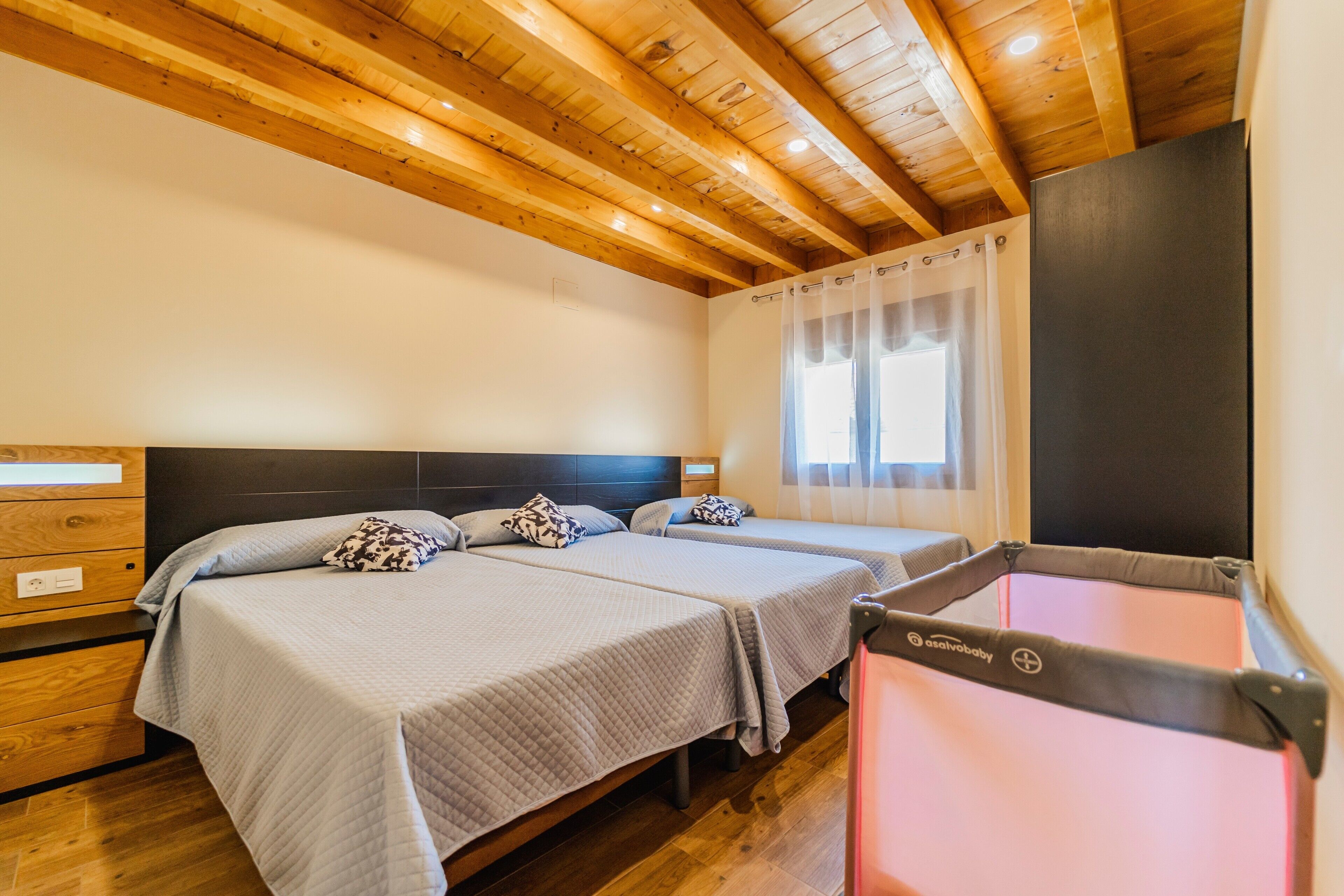 Una habitación de hotel con dos camas, vigas de madera en el techo, una ventana con cortinas y una pared oscura.
