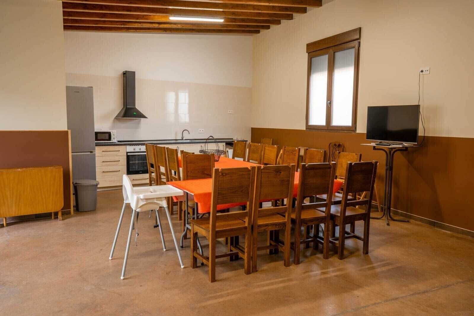 Un coin repas avec une table dressée pour six personnes, des chaises en bois, un téléviseur posé sur un meuble et une cuisine en arrière-plan.