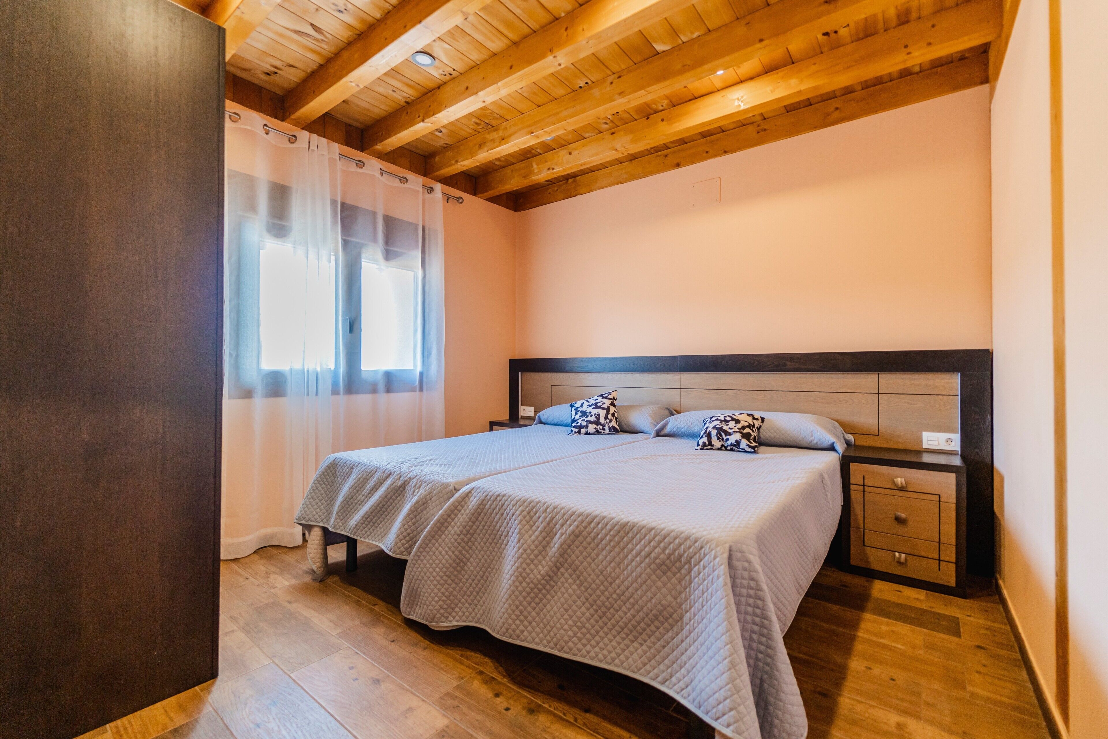 Un dormitorio con techo de madera, una cama con cabecera, una mesita de noche y un ventanal con cortinas.