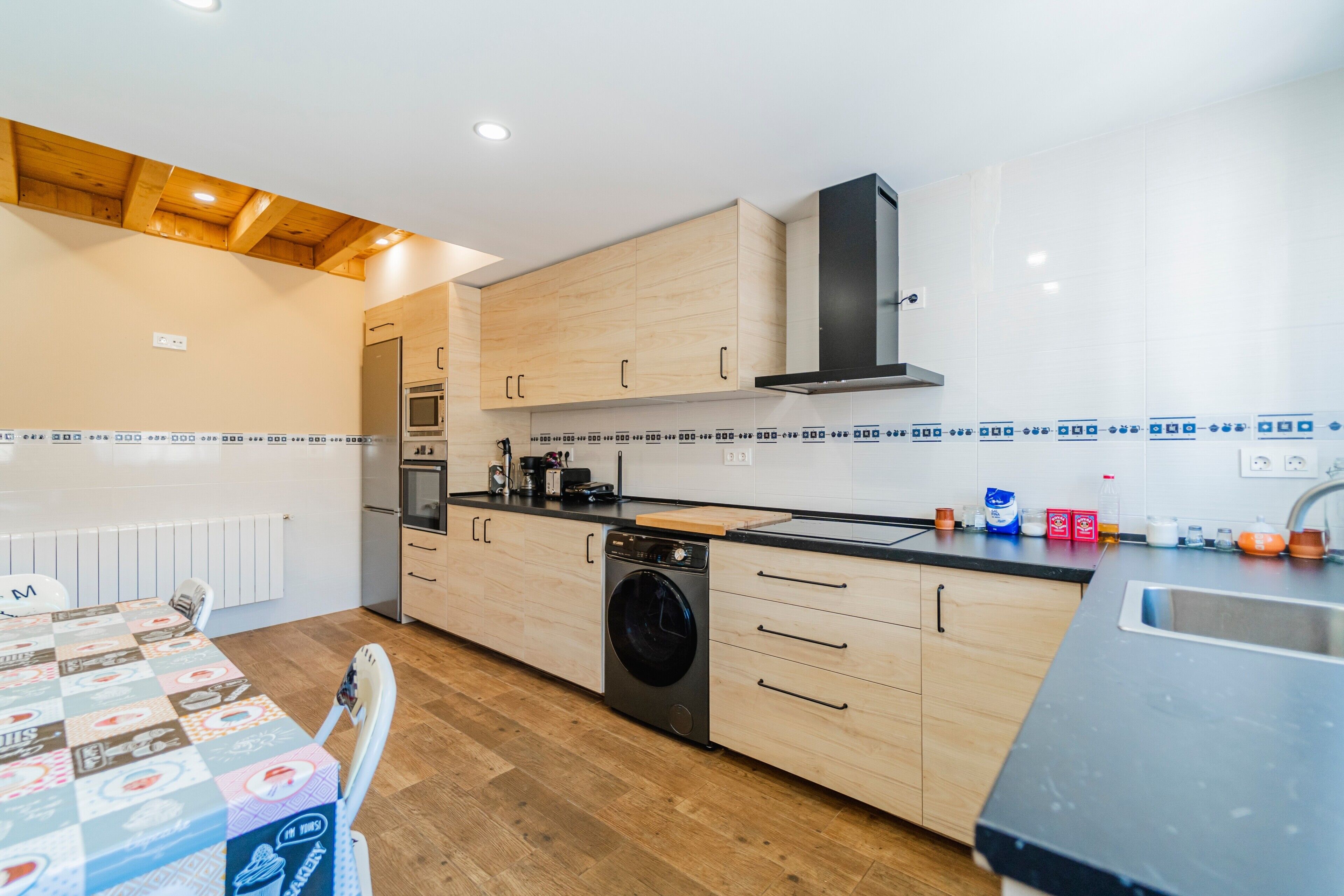 A modern kitchen with wooden cabinets, a built-in oven, a washing machine, and a dining area with a table and chairs.