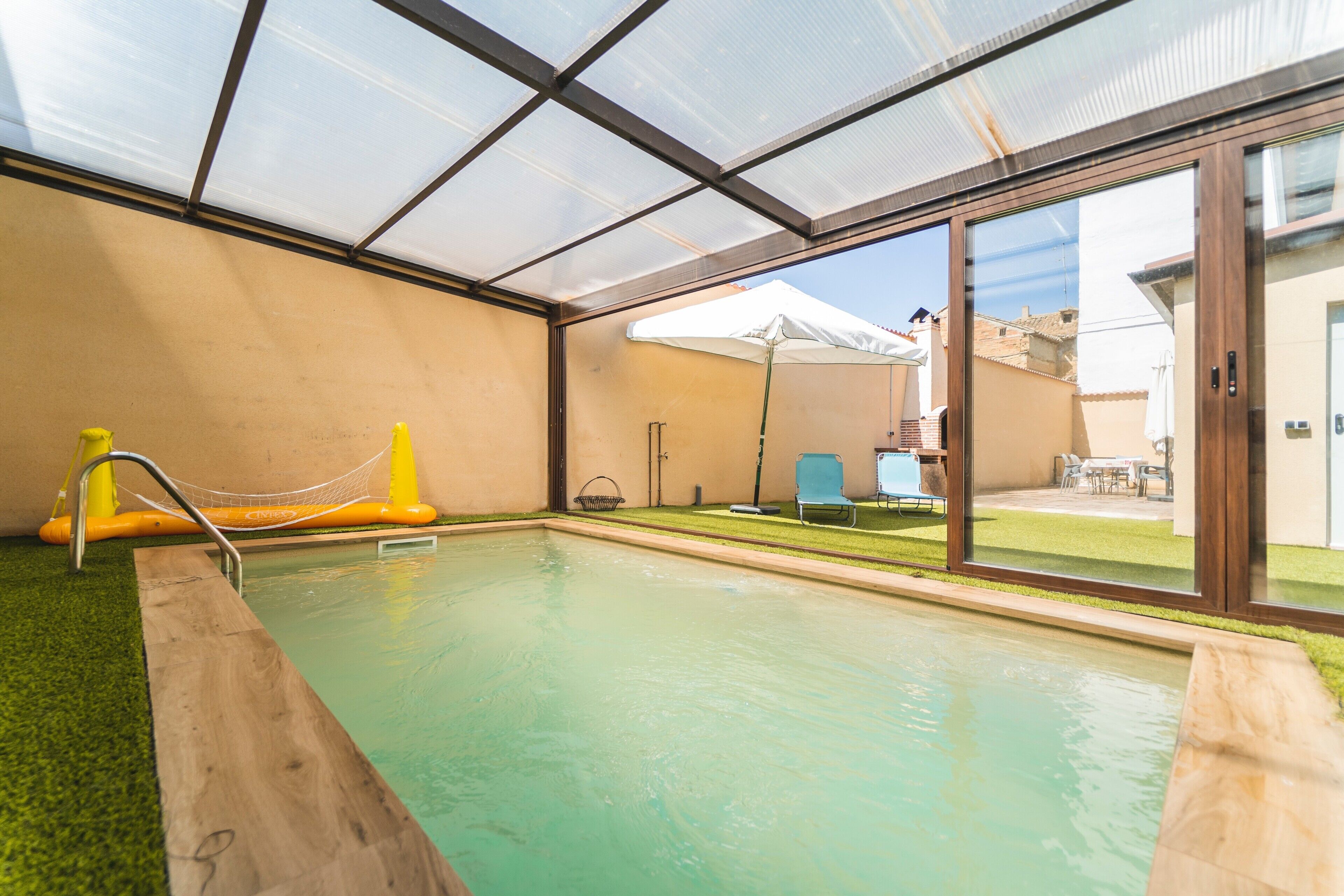 Una piscina interior con techo de vidrio, cubierta de madera y vistas al exterior.