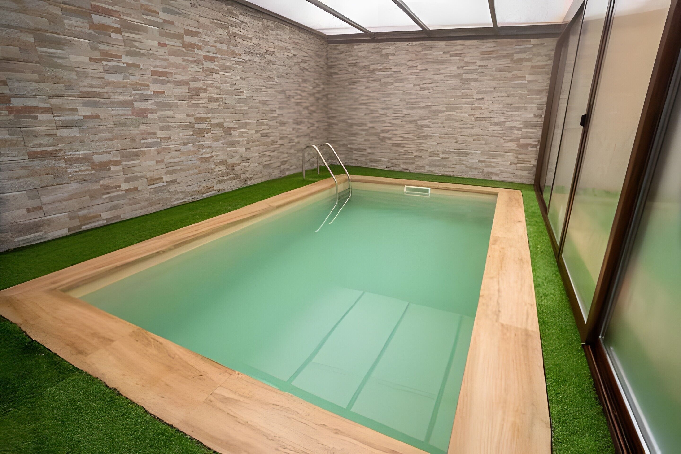 Une piscine intérieure dotée d’une terrasse en bois, entourée de gazon synthétique et de murs en briques.