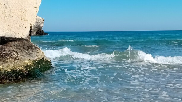Une plage avec une eau cristalline, des vagues et une grande formation rocheuse blanche.