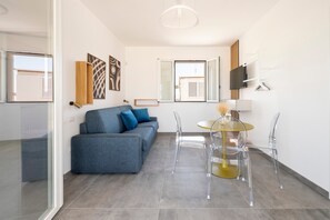 Ein modernes Wohnzimmer mit einer Couch, einem Esstisch mit Stühlen, einem an der Wand befestigten Fernseher und einem Fenster mit Jalousien.